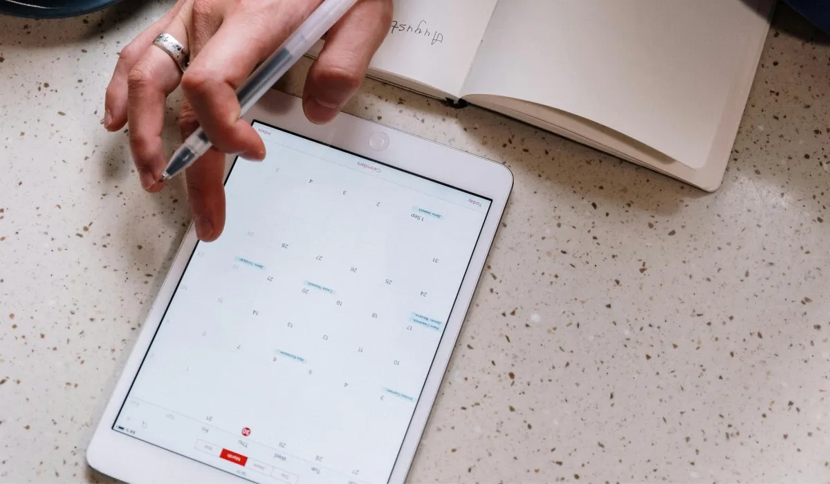 Google Calendar for productivity: a hand holds a pen over a tablet showing a monthly calendar, next to an open notebook on a desk