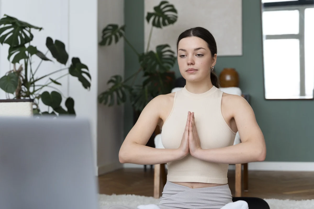 Workplace well-being and physical activity: a woman sits cross-legged with palms together in a calm room with monstera plants and a laptop nearby